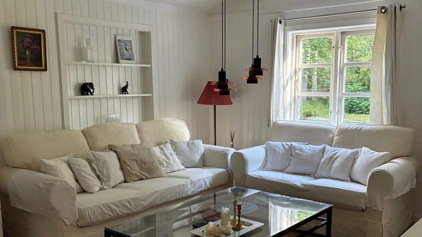 Een woonkamer met twee witte banken, een glazen salontafel, hanglampen, een raam met gordijnen en een witte wand met planken.