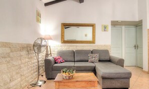 Ein Wohnzimmer mit grauem Sofa, einem Couchtisch mit Früchten, einem Standventilator und einem Spiegel an der Wand.