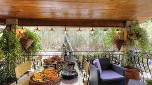 Eine überdachte Terrasse mit Korbmöbeln, einem Tisch mit Speisen und Getränken, hängenden Pflanzen und einem Vogelhäuschen.