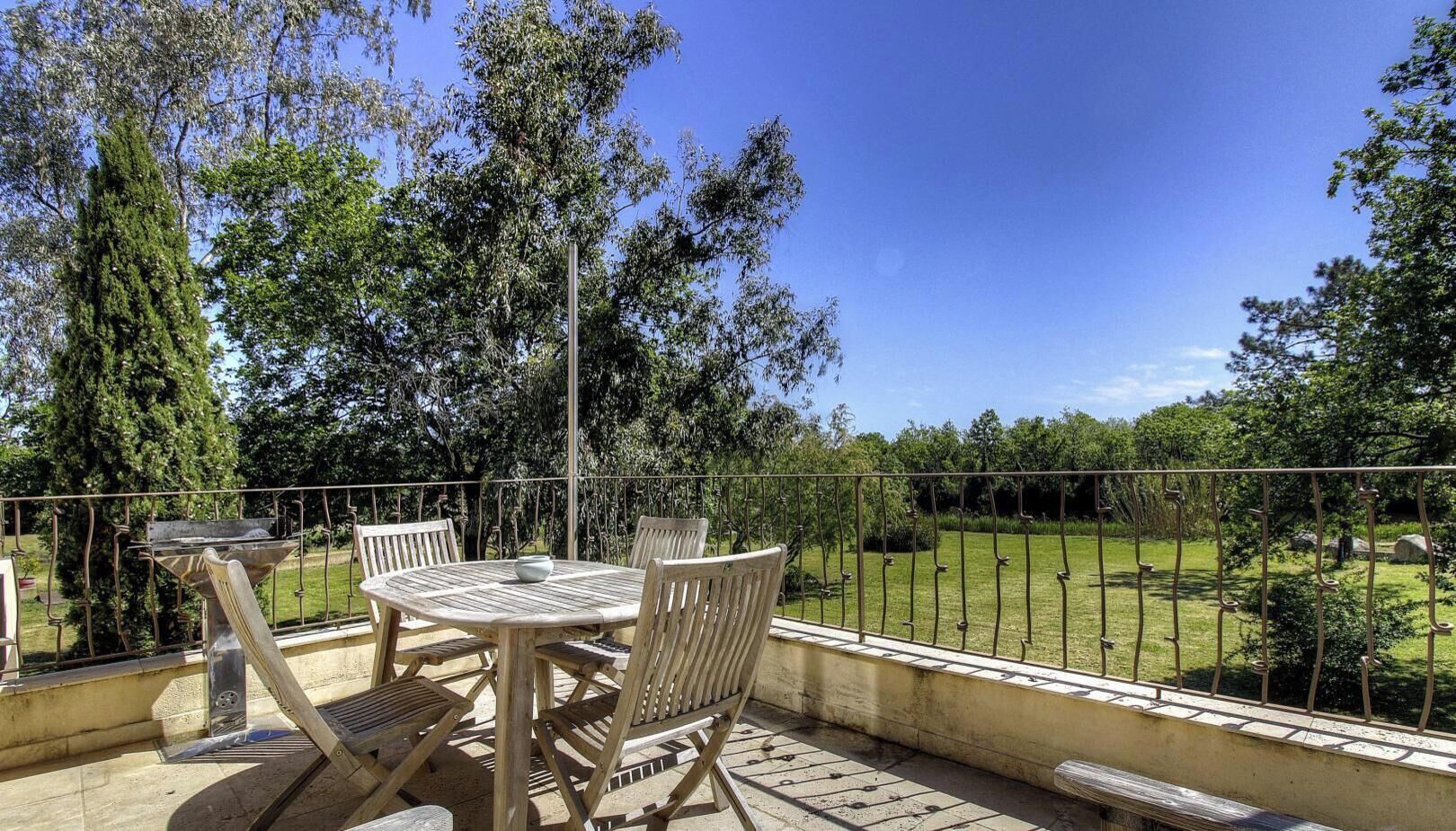 Eine Terrasse im Freien mit einem runden Tisch und Stühlen, einem Grill und Blick auf eine grüne Fläche mit Bäumen.