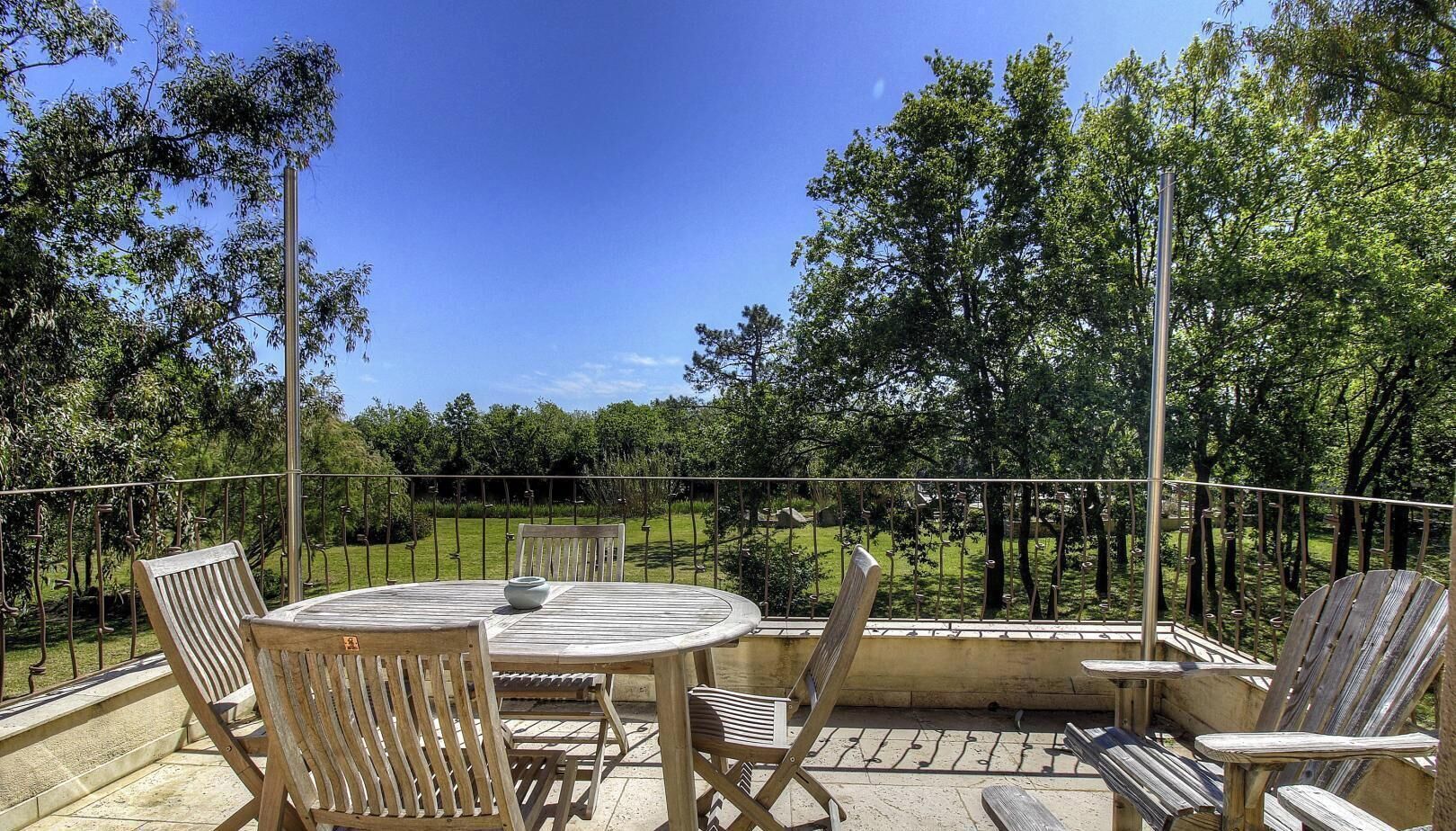 A patio with a round table and chairs, surrounded by a metal fence, overlooking a grassy area with trees.
