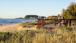 En strand med en träkonstruktion, en sandstrand och ett avlägset kullområde med byggnader.