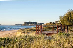 A beach with a wooden structure, a sandy shore, and a distant hill with buildings. - Monkirra One (Miami)