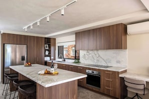 A modern kitchen with a marble countertop, wooden cabinets, a built-in oven, and a large island with bar stools. - Monkirra One (Miami)