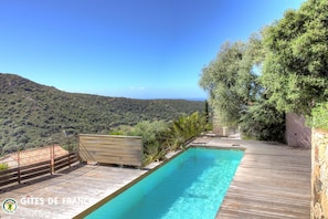 Ein Pool mit Holzterrasse, umgeben von viel Grün und mit Blick auf eine Berglandschaft.