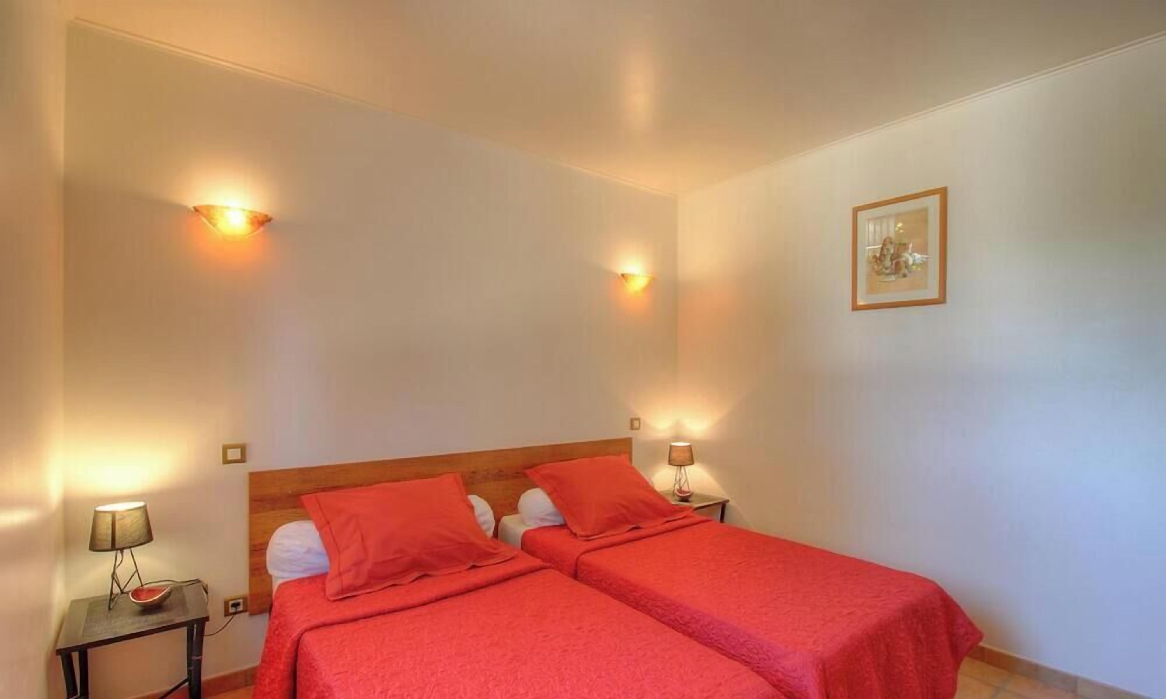 Een kamer met twee bedden, elk met een rode sprei, twee nachtkastjes met lampen, en een inlijsting foto aan de muur.