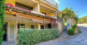 Un edificio a due piani con un portico coperto, un muro in pietra con un cartello e una strada di accesso sterrata.