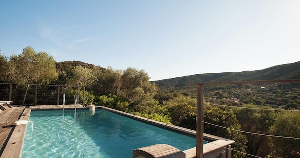 Ein rechteckiger Pool im Freien mit klarem Wasser, umgeben von einer Holzterrasse und Metallgeländern, vor einer Kulisse aus üppigen grünen Bäumen und Hügeln.