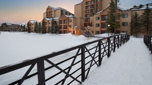Een besneeuwde buitenruimte met een houten hek, een gebouw met meerdere verdiepingen en een skilift in de verte.
