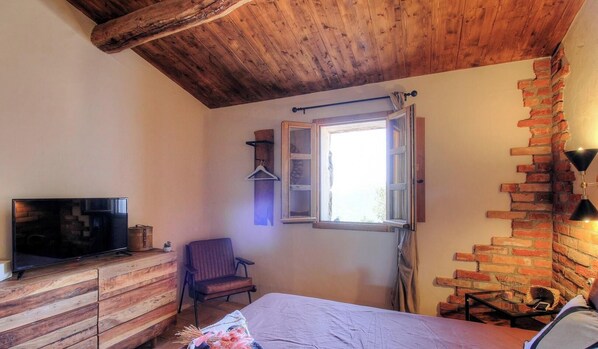 Ein Schlafzimmer mit Holzboden, einem Fernseher auf einer hölzernen Kommode, einem Bett mit weißer Bettdecke, einem Sessel, einem Fenster mit Vorhängen und einer Ziegelwand.