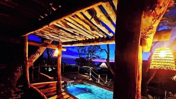 Una terraza de madera con piscina, rodeada de árboles y un cielo nocturno.