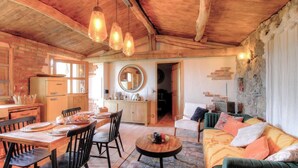 Un interior rústico con vigas de madera a la vista, una zona de comedor con mesa y sillas de madera, un sofá con cojines y una cocina con refrigerador antiguo.