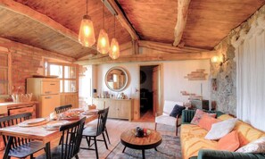 Un interior rústico con vigas de madera a la vista, una zona de comedor con mesa y sillas de madera, un sofá con cojines y una cocina con refrigerador antiguo.