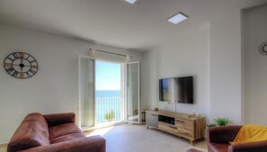 Una sala de estar con televisor, sofá, una mesa de centro y un ventanal corredizo que da a un balcón con vistas.