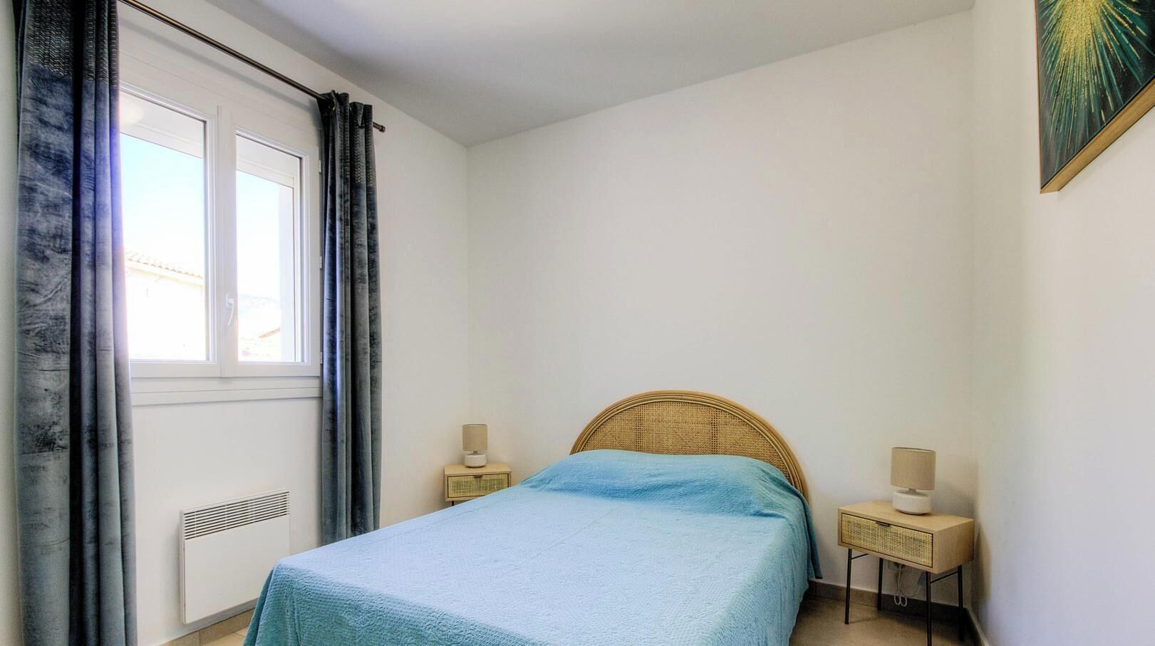 A bedroom with a bed, two bedside tables, a window with curtains, and a wall-mounted artwork.