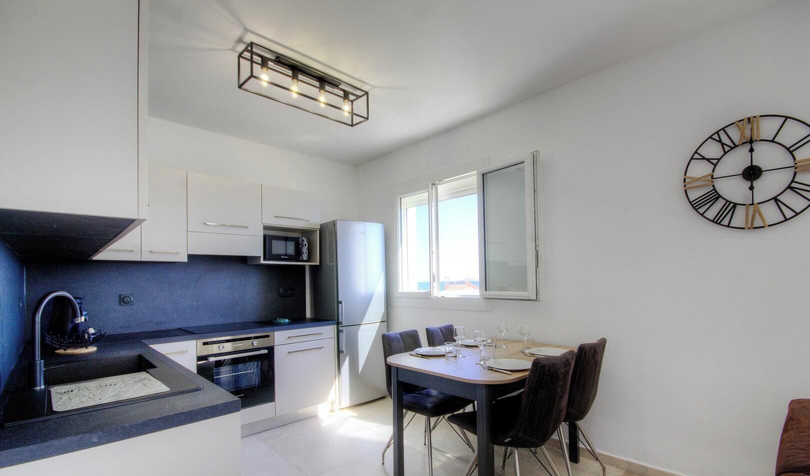 A modern kitchen with a dining area, a microwave, a refrigerator, and a wall clock.