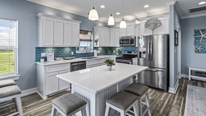 A modern kitchen with white cabinetry, a central island, bar stools, stainless steel appliances, and a view of the outdoors. - Beach Access~Barefoot Bliss~Chef Kitchen~Family (Bolivar Peninsula)