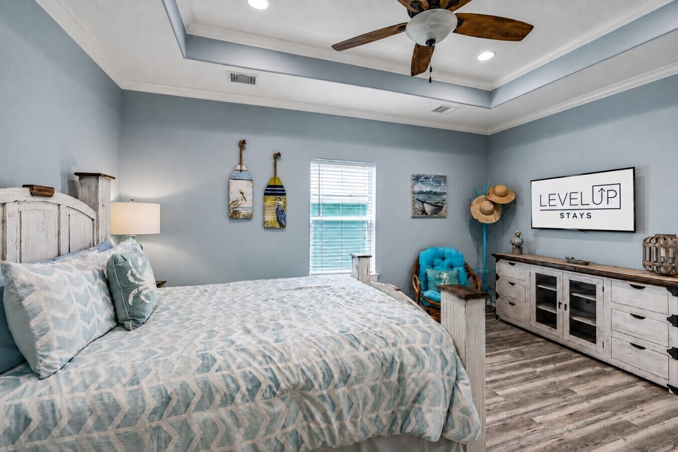 A bedroom with a bed, a ceiling fan, a window with blinds, a chair, a dresser, and wall art.