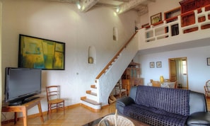 Un saló amb televisió, sofà, cadires, una escala i zona de menjador.
