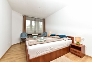 Ein Schlafzimmer mit einem hölzernen Bett, einem Schreibtisch, zwei Stühlen, einem Nachttisch mit Lampe und einem Fenster mit Vorhängen.