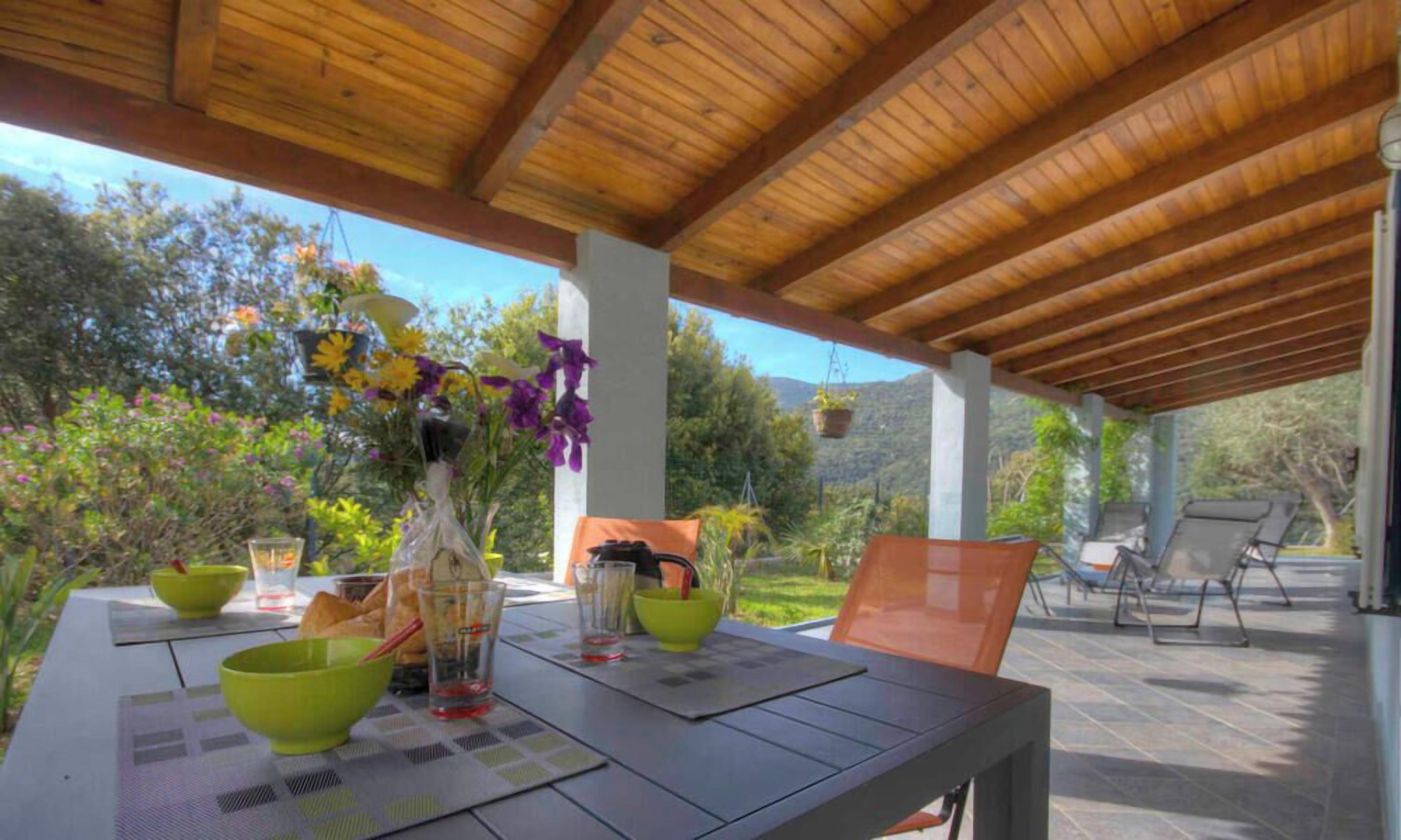 An outdoor patio with a wooden ceiling, a table set with bowls, glasses, and a bottle, surrounded by chairs and plants.