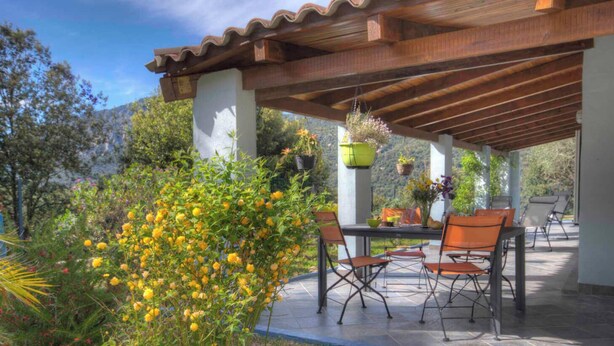Een terras buiten met een houten pergola, een tafel met stoelen en hangende bloemenplanters.
