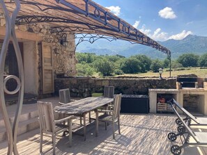 Eine Terrasse im Freien mit einer Steinmauer, einem Holztisch und Stühlen, einem Grill und einer Metallpergola.
