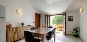 A dining area with a wooden table and chairs, a cabinet with decorative items, and a view of the outdoors through an open door. - Casa Stella Marina - SAINT FLORENT (SAINT FLORENT)