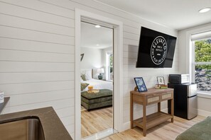 Een moderne hotelkamer met een bed, een klein tafeltje met een fotolijst, een aan de muur bevestigde televisie en een raam met uitzicht op bomen.