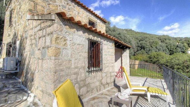 Un edificio in pietra con un balcone, una finestra con persiane e vista su una collina boscosa.