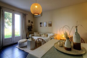 Ein Wohnzimmer mit einer Couch, einem Esstisch mit Vasen, einer Hängelampe und einem großen Fenster mit Vorhängen.