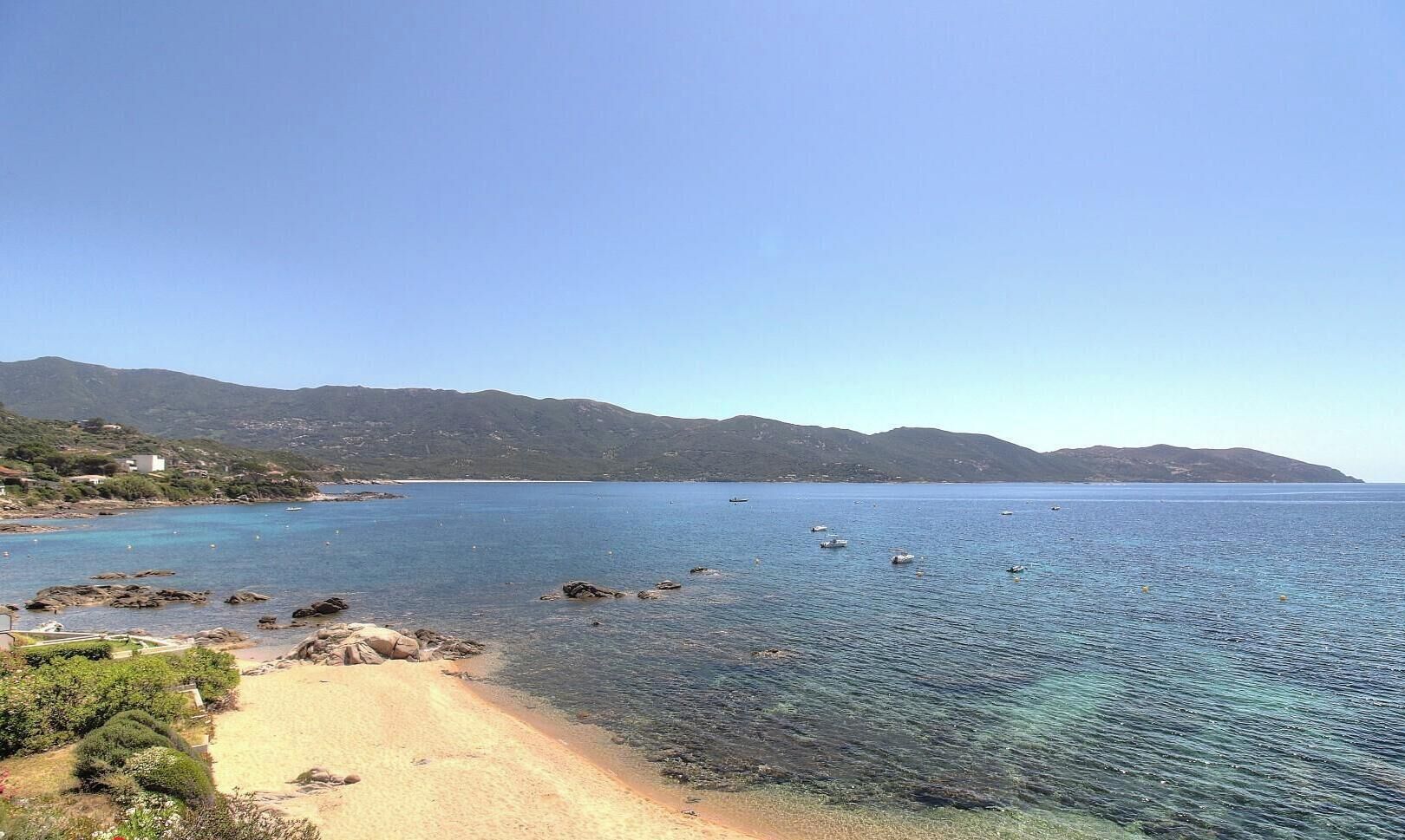 A coastal landscape with clear water, a sandy beach, and distant mountains.