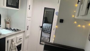 Eine kleine Küche mit weißen Schränken, einem Herd und einer kompakten Arbeitsfläche. Ein Spiegel spiegelt den Raum wider, und durch eine offene Tür ist ein Bett zu sehen.