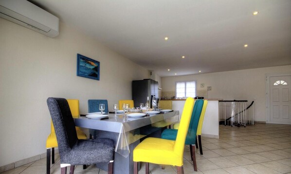A dining area with a table set for four, chairs with yellow and blue upholstery, a kitchen in the background, and a staircase leading to another level. - Villa Alivu - GALERIA (GALERIA)