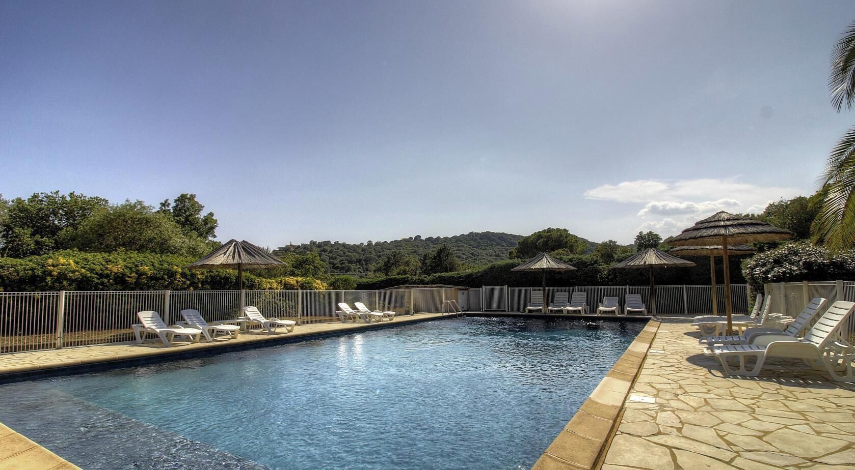 Una piscina con lettini e ombrelloni, circondata da una recinzione e vegetazione.