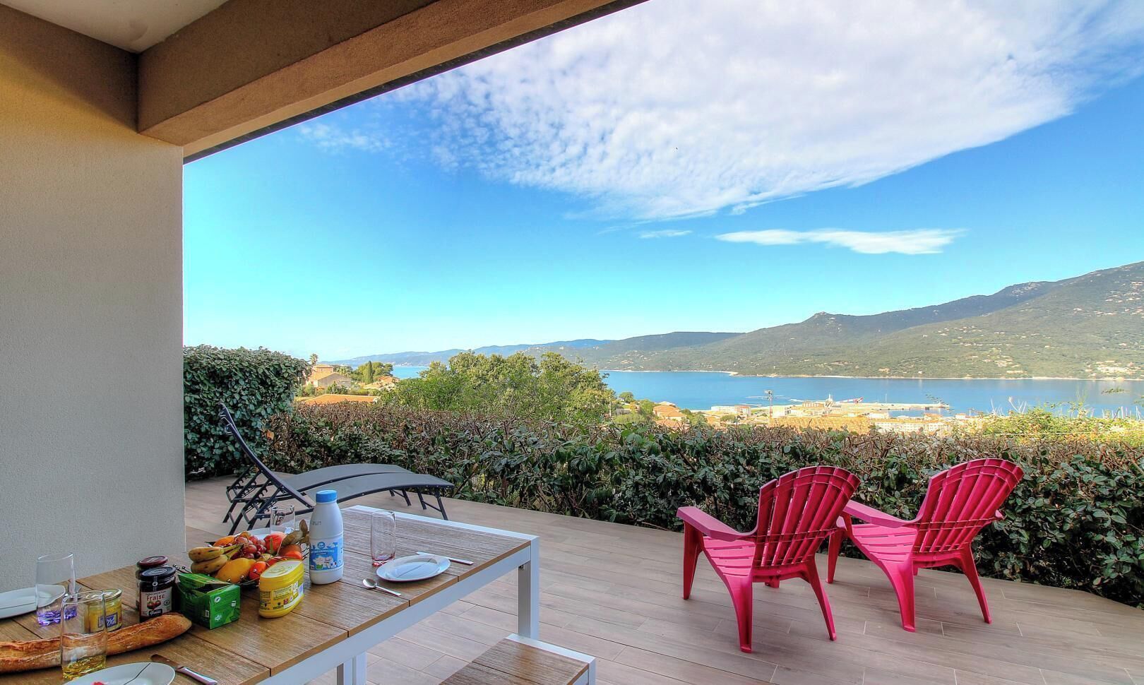 Ein Balkon mit Frühstückstisch, zwei roten Stühlen und einem malerischen Blick auf See und Berge.