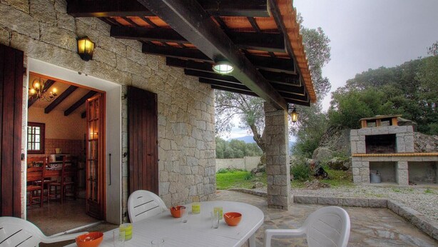 A stone building with a dining area featuring a table and chairs, a kitchen visible through an open door, and a stone structure with a chimney in the background. - I caseddu d'Ulmetu1 - OLMETO (OLMETO)