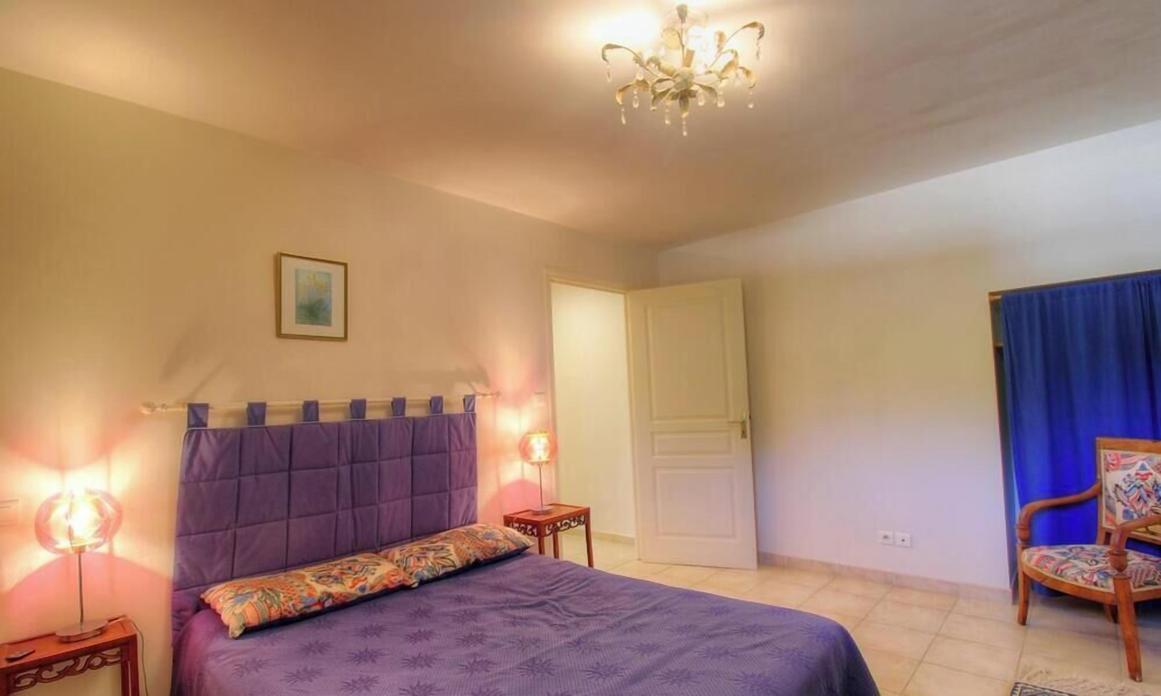 A bedroom with a bed, bedside lamps, a chandelier, a door, and a chair.