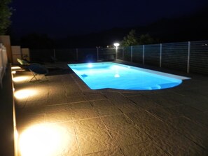 Ein Pool mit gefliestem Deck, umgeben von einem Zaun und beleuchtet durch Lichter.