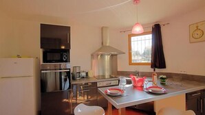 Et lille køkken med mikroovn, komfur og vask, et spisebord med tallerkener og en flaske samt et vindue med gardiner.