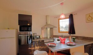 A compact kitchen with a microwave, stove, and sink, a dining table with plates and a bottle, and a window with curtains. - Le Lilas - SOTTA - Gîte (SOTTA)