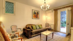 Een woonkamer met een bank, stoelen, een glazen salontafel, een kroonluchter en een deur naar een buitengebied.