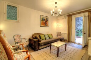 Ein Wohnzimmer mit einer Couch, Sesseln, einem Glastisch, einer Deckenleuchte und einer Tür, die zu einem Außenbereich führt.