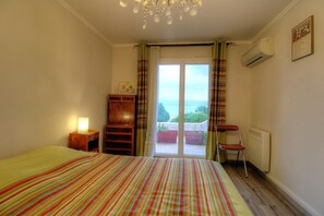 Ein Schlafzimmer mit Bett, Nachttisch, Holzkommode, Stuhl und Heizkörper, sowie Blick ins Freie.