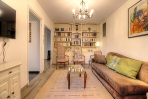 Ein Wohnzimmer mit Sofa, Sesseln, Couchtisch, Bücherregal, Fernseher und einer Deckenleuchte.