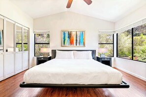 Ein Schlafzimmer mit einem Bett, zwei Nachttischen, einem Deckenventilator, einem großen Fenster und einem Gemälde an der Wand.