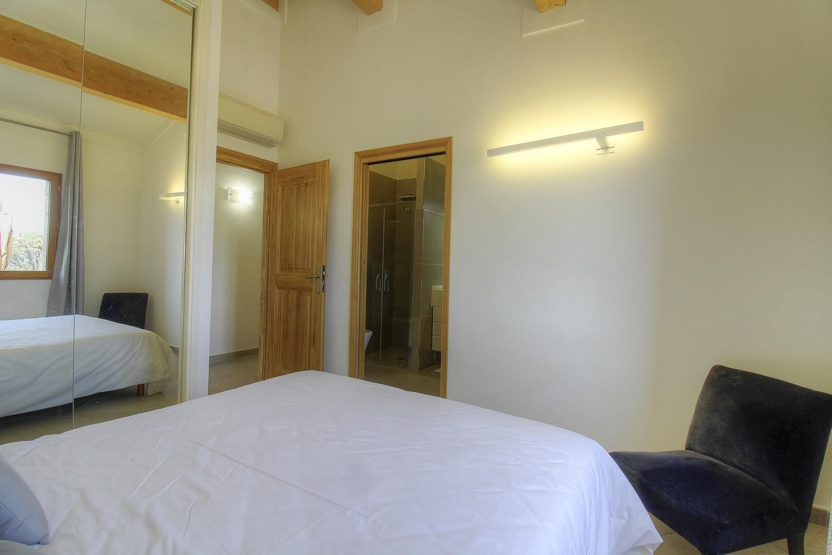 A bedroom with a bed, a chair, a wooden door, and a shower area.