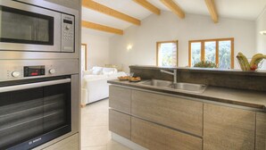 A modern kitchen with a Bosch microwave, oven, and a built-in sink. There is a dining area with a white sofa and a window with a view of greenery.