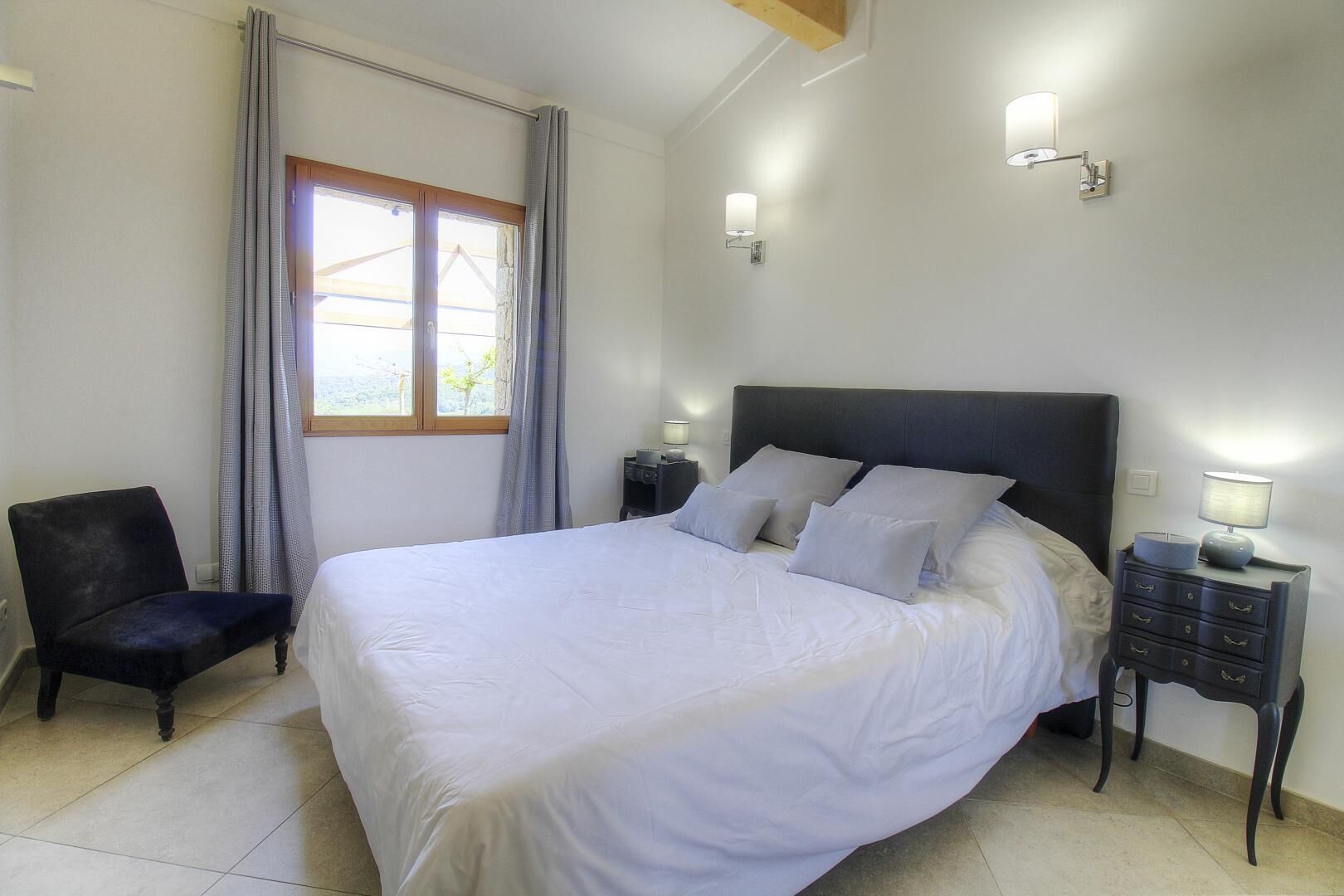 Une chambre à coucher avec un grand lit, une chaise, une table de chevet et une fenêtre avec des rideaux.
