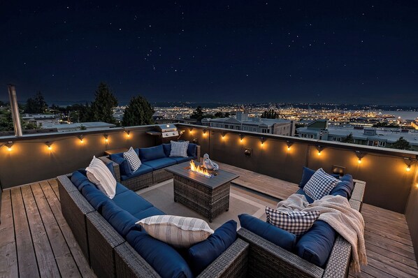 A rooftop terrace with a fire table, blue sofas, and string lights against a night sky.